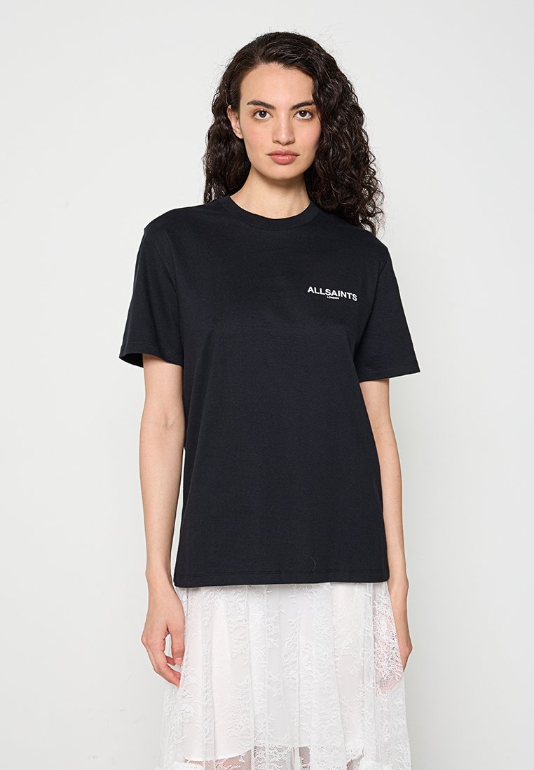 Frau mit lockigem Haar, die ein schwarzes AllSaints-Logo-T-Shirt und einen weißen Spitzenrock trägt, steht vor einem schlichten hellen Hintergrund.