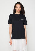 Frau mit lockigem Haar, die ein schwarzes AllSaints-Logo-T-Shirt und einen weißen Spitzenrock trägt, steht vor einem schlichten hellen Hintergrund.