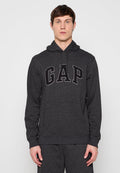 Mann trägt dunkelgrauen Kapuzenpullover mit großem "GAP"-Logo auf der Brust, steht vor einfachem hellem Hintergrund.
