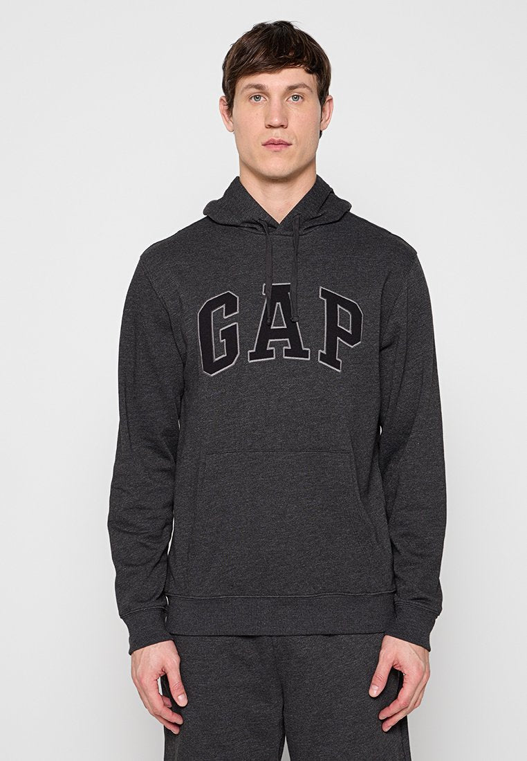 Mann trägt dunkelgrauen Kapuzenpullover mit großem "GAP"-Logo auf der Brust, steht vor einfachem hellem Hintergrund.