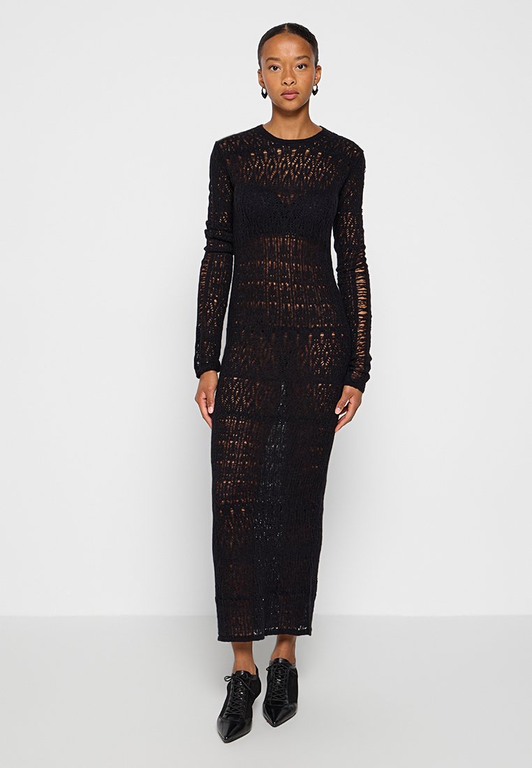 Schwarzes langes Strickkleid mit strukturiertem, durchsichtigen Design, gerader Silhouette und Rundhalsausschnitt. Getragen mit schwarzen spitzen Schuhen.
