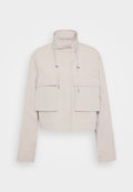 Gekürzte beige Jacke aus leichtem Stoff. Verfügt über einen hohen Kragen, zwei aufgesetzte Taschen vorne und verstellbare Kordeln am Hals.