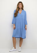 Helles blaues, knielanges Kleid mit V-Ausschnitt, dreiviertellangen Glockenärmeln und gestuftem Rock aus weichem Stoff, getragen mit weißen Sneakers.