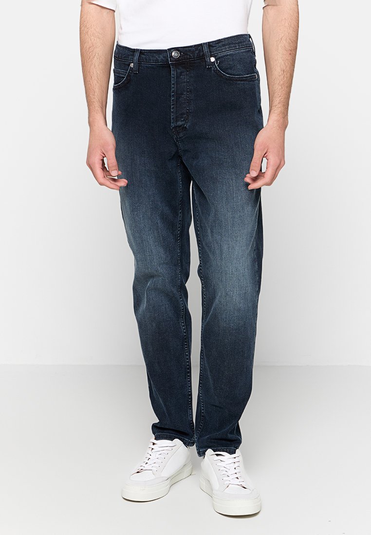 Dunkelblaue Jeans aus Denim mit geradem Schnitt, ausgestattet mit fünf Taschen, Gürtelschlaufen und einem leichten Verblassungseffekt. Kombiniert mit weißen Turnschuhen.