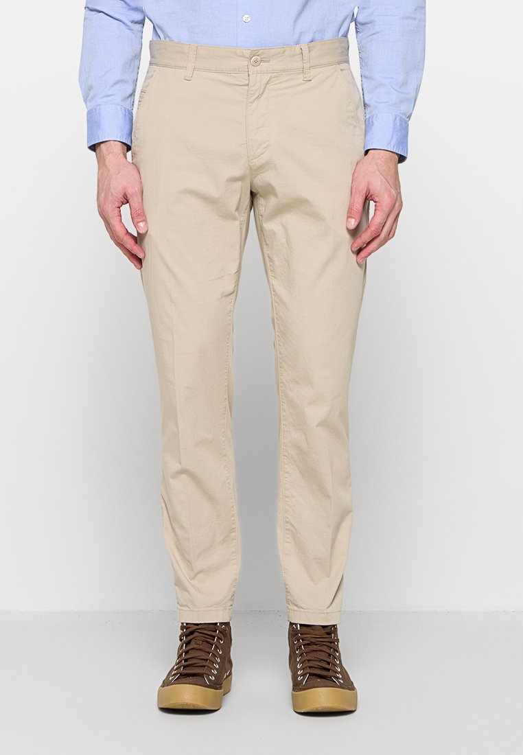 Mann trägt beigefarbene Slim-Fit-Hose, hellblaues Langarmhemd und braune High-Top-Sneaker, steht vor einem schlichten weißen Hintergrund.
