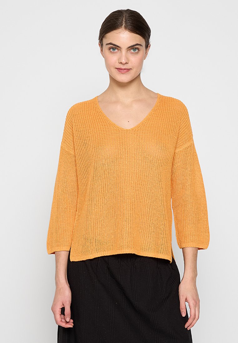 V-Ausschnitt, locker sitzender Pullover in einer leuchtend orangefarbenen Strickstruktur, mit Dreiviertelärmeln und geradem Saum. Getragen mit einem schwarzen Rock.