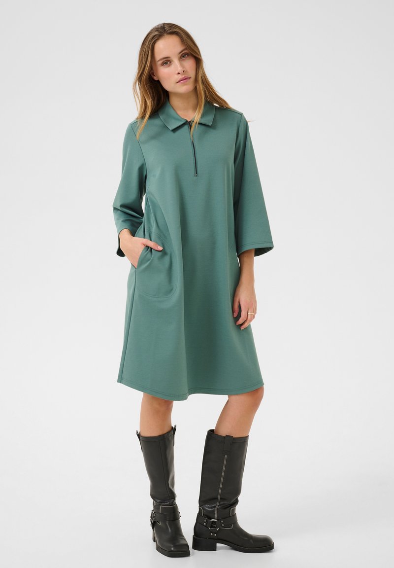 Grünes Polo-Kleid mit Reißverschluss am Ausschnitt, dreiviertellangen Ärmeln und Seitentaschen, kombiniert mit schwarzen, kniehohen Stiefeln und Schnallendetails.