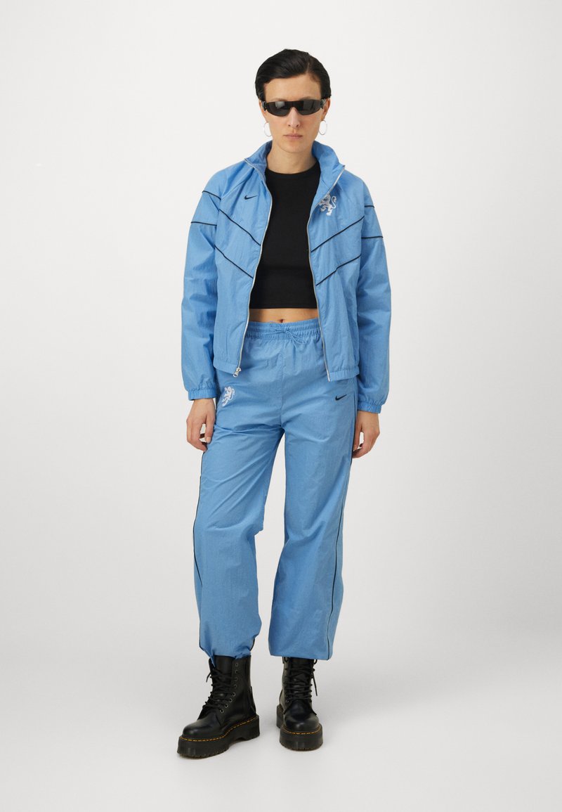 Helle blaue Trainingsanzug mit schwarzen Akzenten, bestehend aus einer Reißverschlussjacke und Hose mit elastischem Bund, kombiniert mit schwarzen Kampfstiefeln und Sonnenbrille.