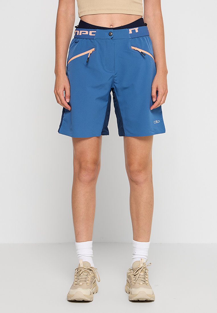 Frau in blauen Outdoor-Shorts mit pfirsichfarbenen Reißverschlusstaschen, weißen Socken und beigen Wanderschuhen, die vor einem schlichten Hintergrund steht.