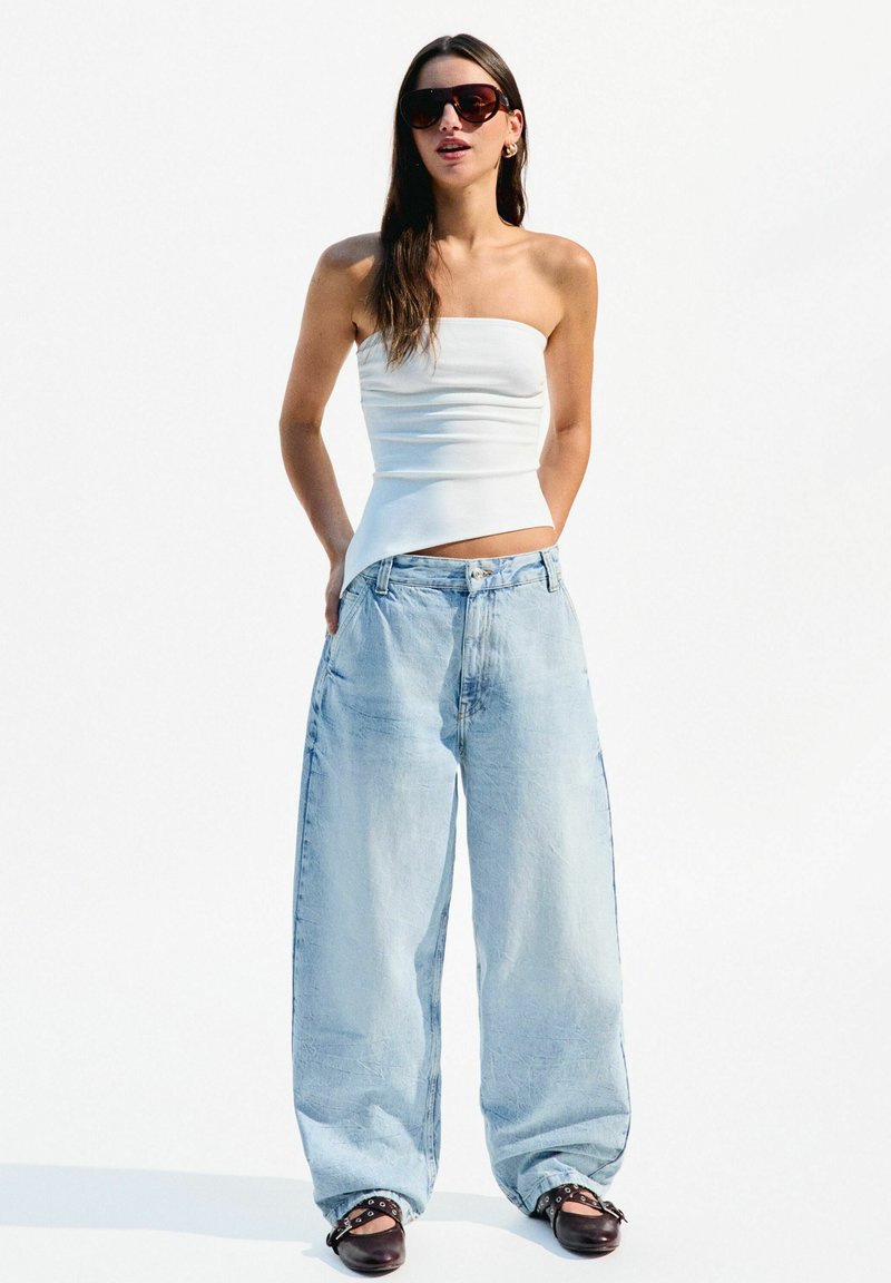 Weißes trägerloses Top, lockere hellblaue Jeans mit ausgewaschenem Finish und braune High Heels. Die Accessoires umfassen übergroße Sonnenbrillen.