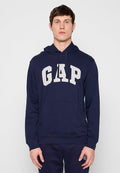 Junger Mann, der einen dunkelblauen Hoodie mit dem großen weißen "GAP"-Logo trägt, steht vor einem schlichten hellgrauen Hintergrund.