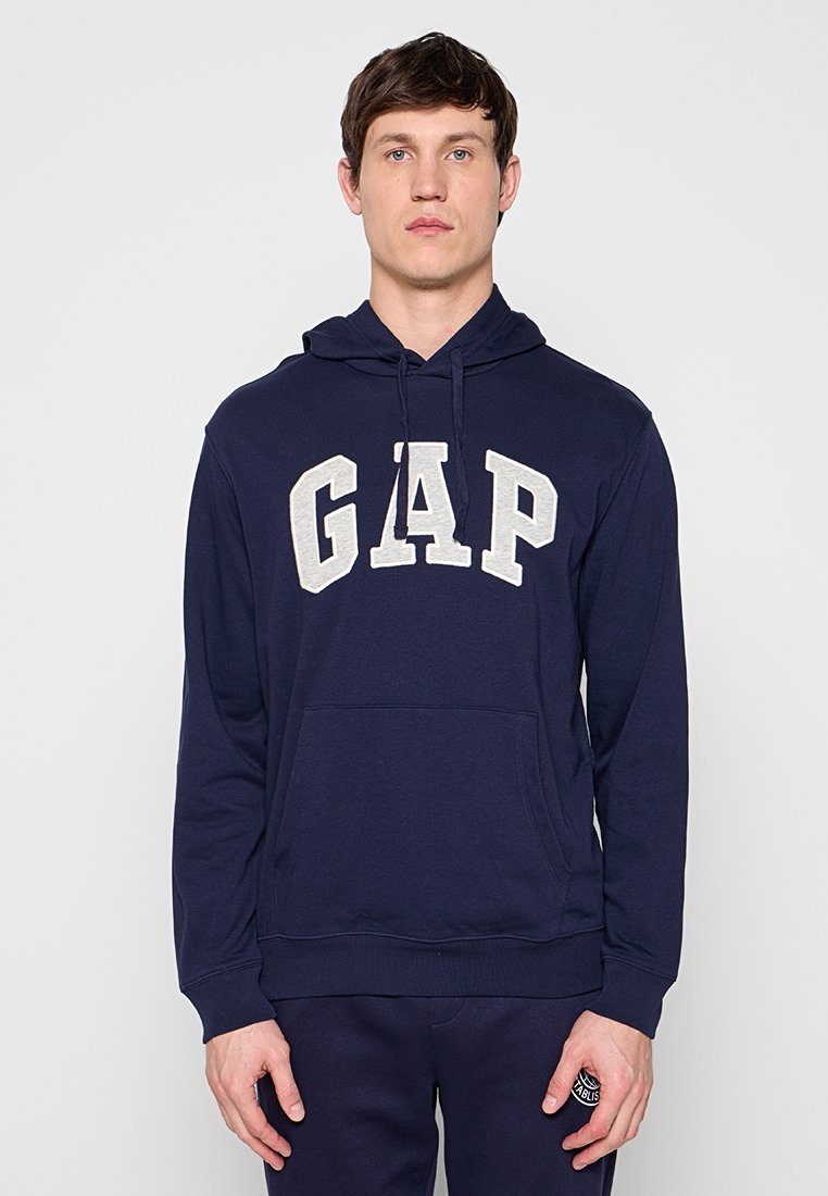 Junger Mann, der einen dunkelblauen Hoodie mit dem großen weißen "GAP"-Logo trägt, steht vor einem schlichten hellgrauen Hintergrund.