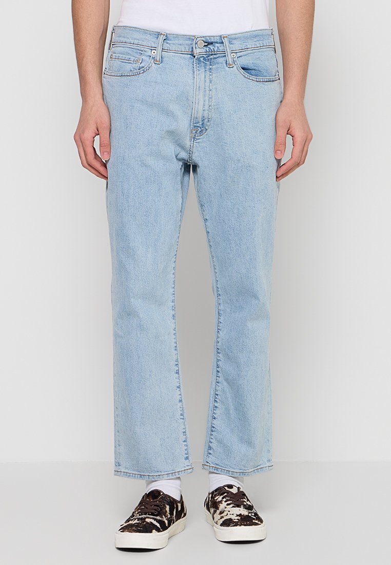 Hellblaue Jeans mit geradem Bein, fünf Taschen und subtilen Verwaschungen. Getragen mit gemusterten braunen Sneakern und weißen Socken.