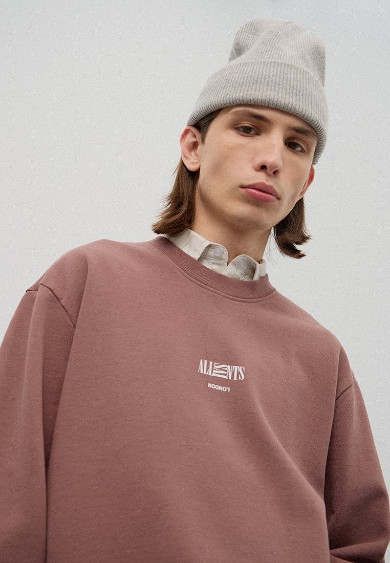 Brauner Hoodie mit weißem Logo auf der Brust, runden Halsausschnitt und langen Ärmeln. Tragen mit einer hellgrauen gerippten Mütze. Subtile Stofftextur.