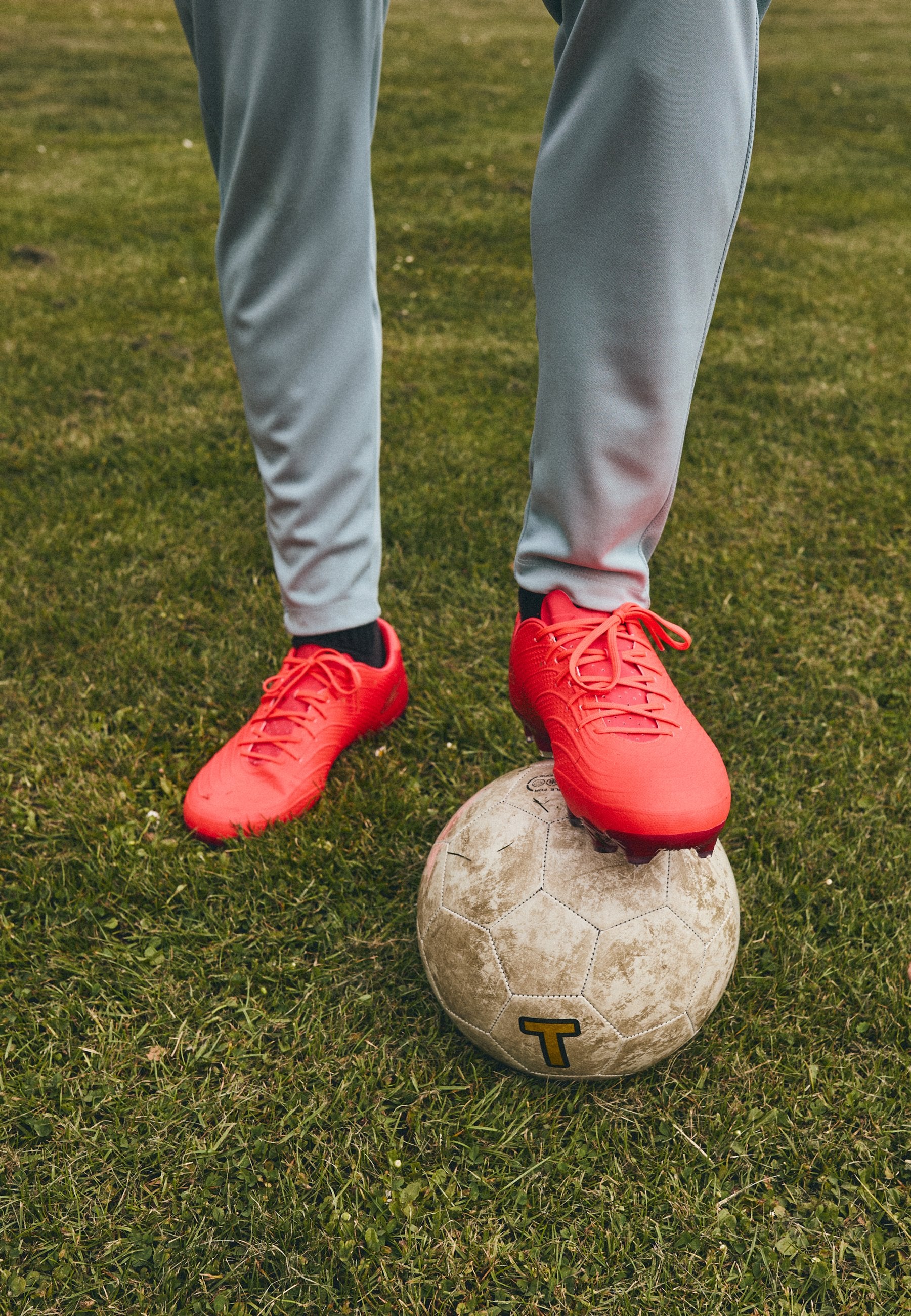 Rote Fußballschuhe mit strukturierten Oberflächen und Schnürsenkeln, stehen auf einem abgenutzten grauen Fußball, auf Gras mit sichtbaren Halmen.
