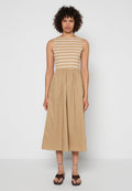 Frau mit lockigem Haar, die ein ärmelloses Kleid in Beige und Weiß mit Streifen und geraffter Taille sowie braune, offene High Heels trägt.