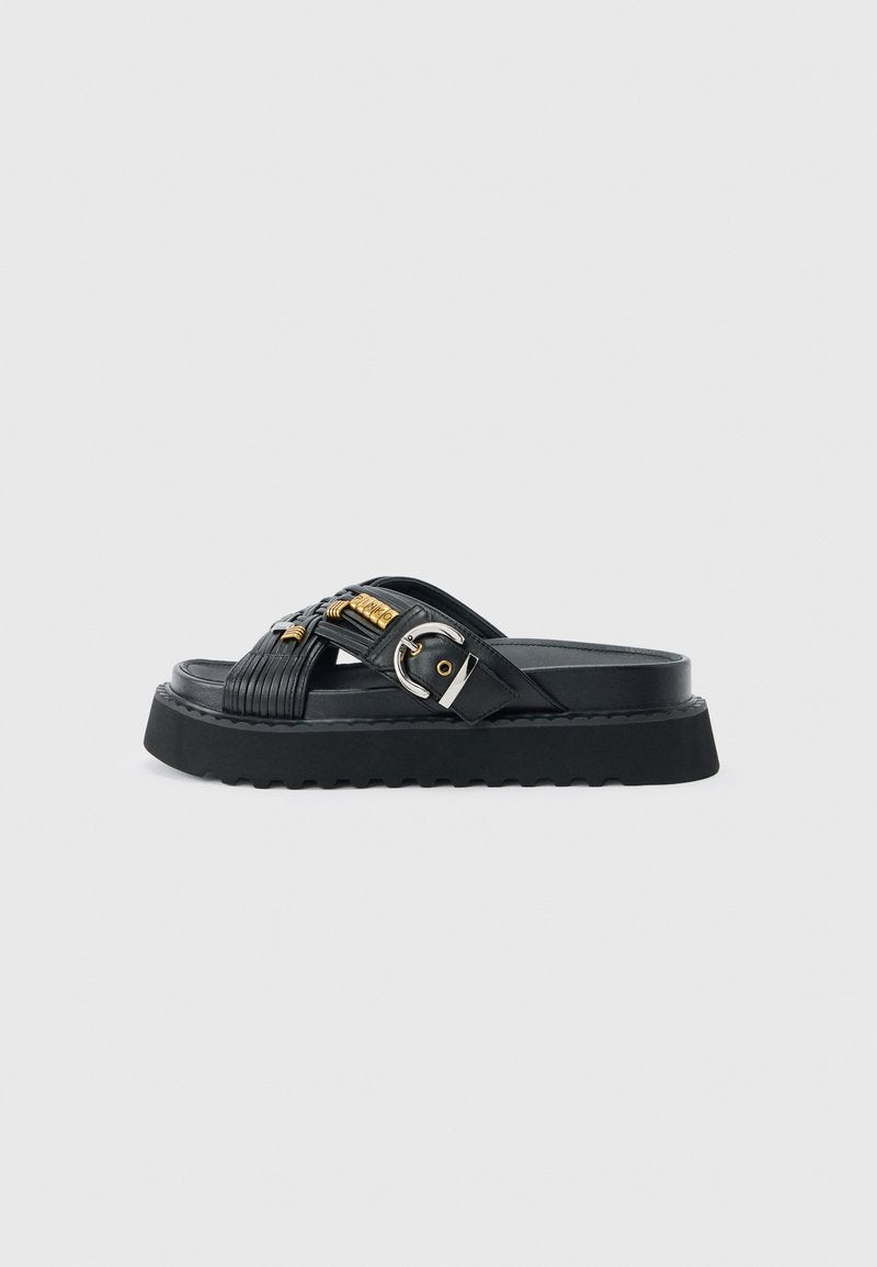 Schwarze Slidesandalette mit dicker Sohle, überkreuzten Riemen mit goldenen Akzenten und einer silbernen Schnalle auf grauem Hintergrund.