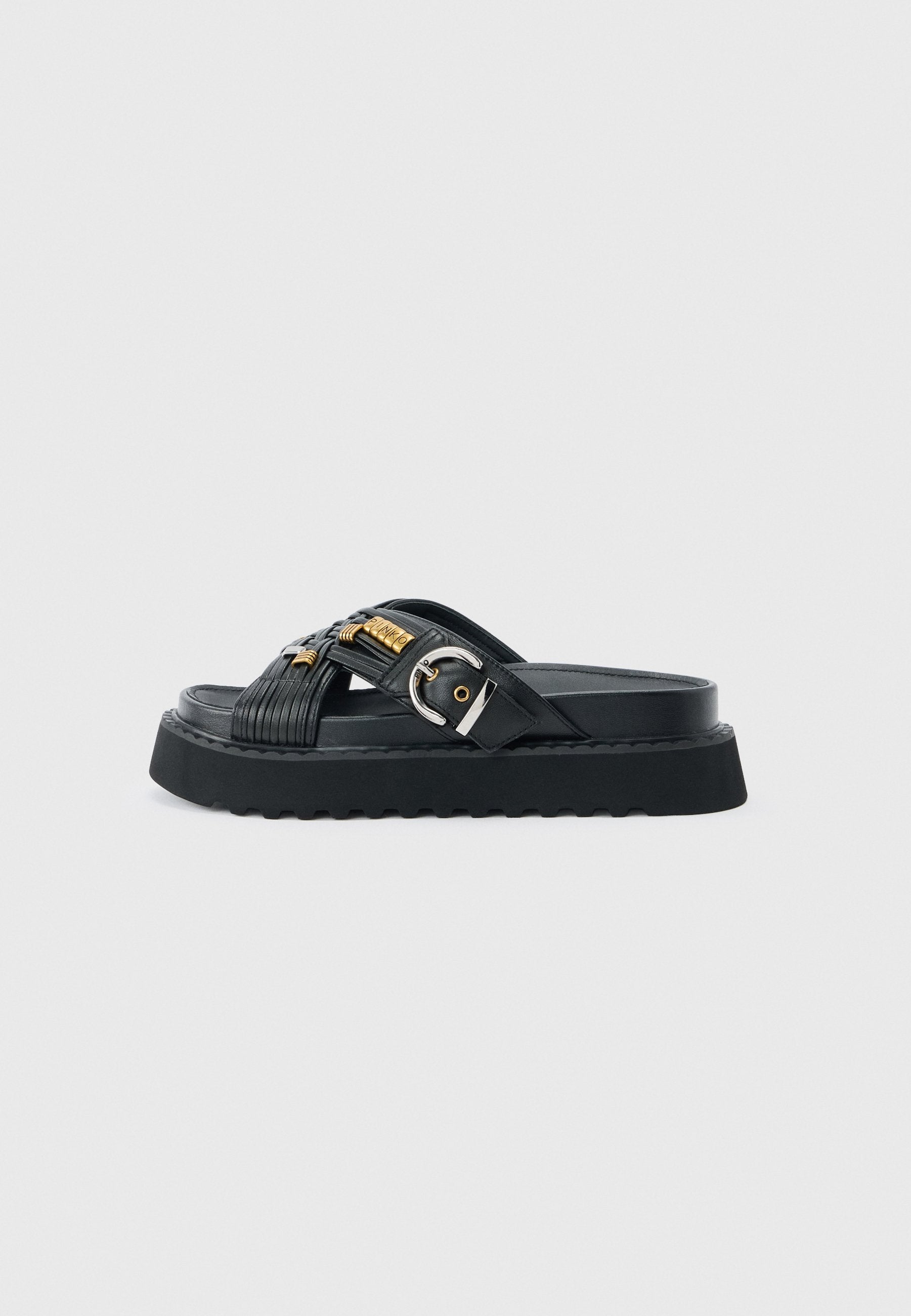 Schwarze Slidesandalette mit dicker Sohle, überkreuzten Riemen mit goldenen Akzenten und einer silbernen Schnalle auf grauem Hintergrund.