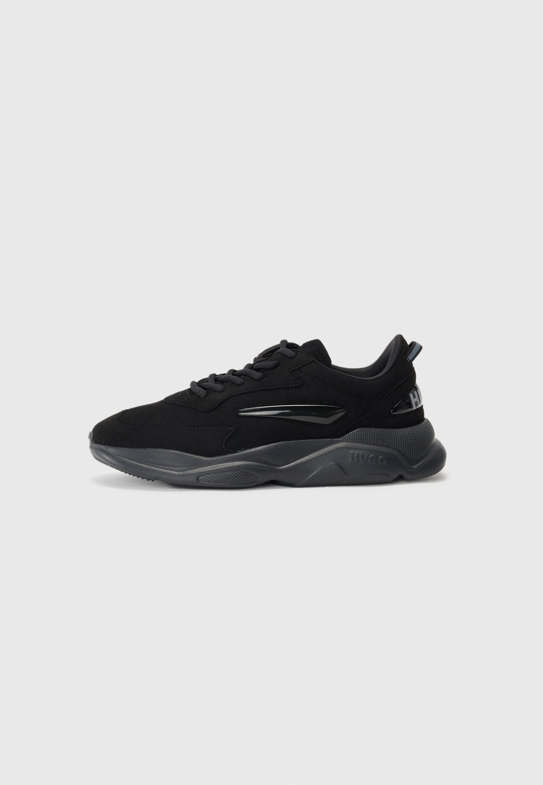 Schwarze Sneakers mit einem Obermaterial aus Wildleder, ausgestattet mit einem glänzenden schwarzen Akzent, einer strukturierten grauen Sohle und einer Schnürdesign. Enthält Branding an der Ferse.