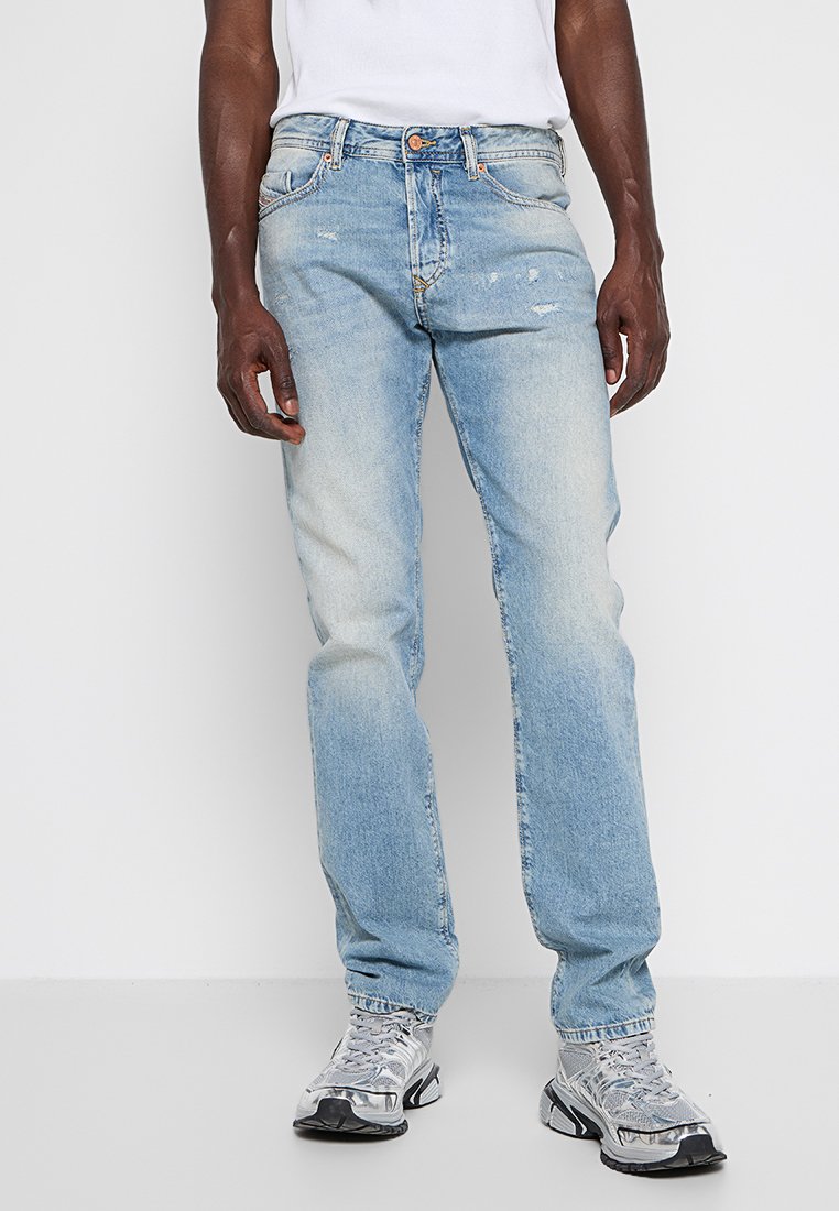 Hellblaue Denim-Jeans mit geradem Schnitt, abgerissenen Patches und Fünf-Taschen-Design. Kombiniert mit grauen Sportschuhen.