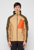 Mann trägt beige und olivgrüne Windjacke mit vordertasche in orange auf der Brust, steht vor einfachem hellem Hintergrund.