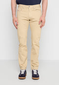 Ein Mann trägt beige Slim-Fit-Hosen und marineblaue Sneaker mit weißen Schnürsenkeln und steht vor einem einfarbigen hellen Hintergrund.