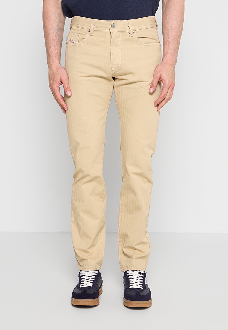 Ein Mann trägt beige Slim-Fit-Hosen und marineblaue Sneaker mit weißen Schnürsenkeln und steht vor einem einfarbigen hellen Hintergrund.