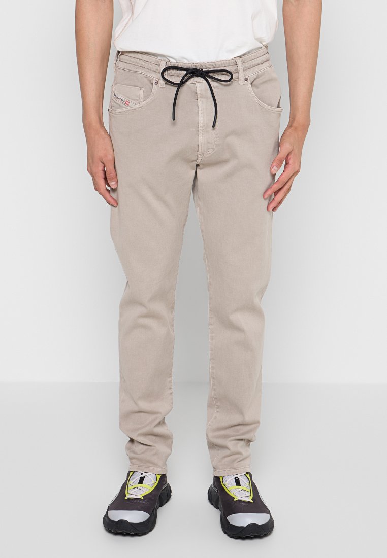 Beige Baumwollhosen mit einem Kordelzugbund, zwei frontalen Taschen und einem dezenten Logo-Patch. Getragen mit schwarz-grauen Turnschuhen.