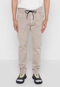 Beige Baumwollhosen mit einem Kordelzugbund, zwei frontalen Taschen und einem dezenten Logo-Patch. Getragen mit schwarz-grauen Turnschuhen.