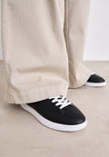 Schwarze Ledersneaker mit weißen Schnürsenkeln und einer weißen Gummisohle, kombiniert mit lockeren beigen Hosen, auf einer hellen strukturierten Oberfläche.