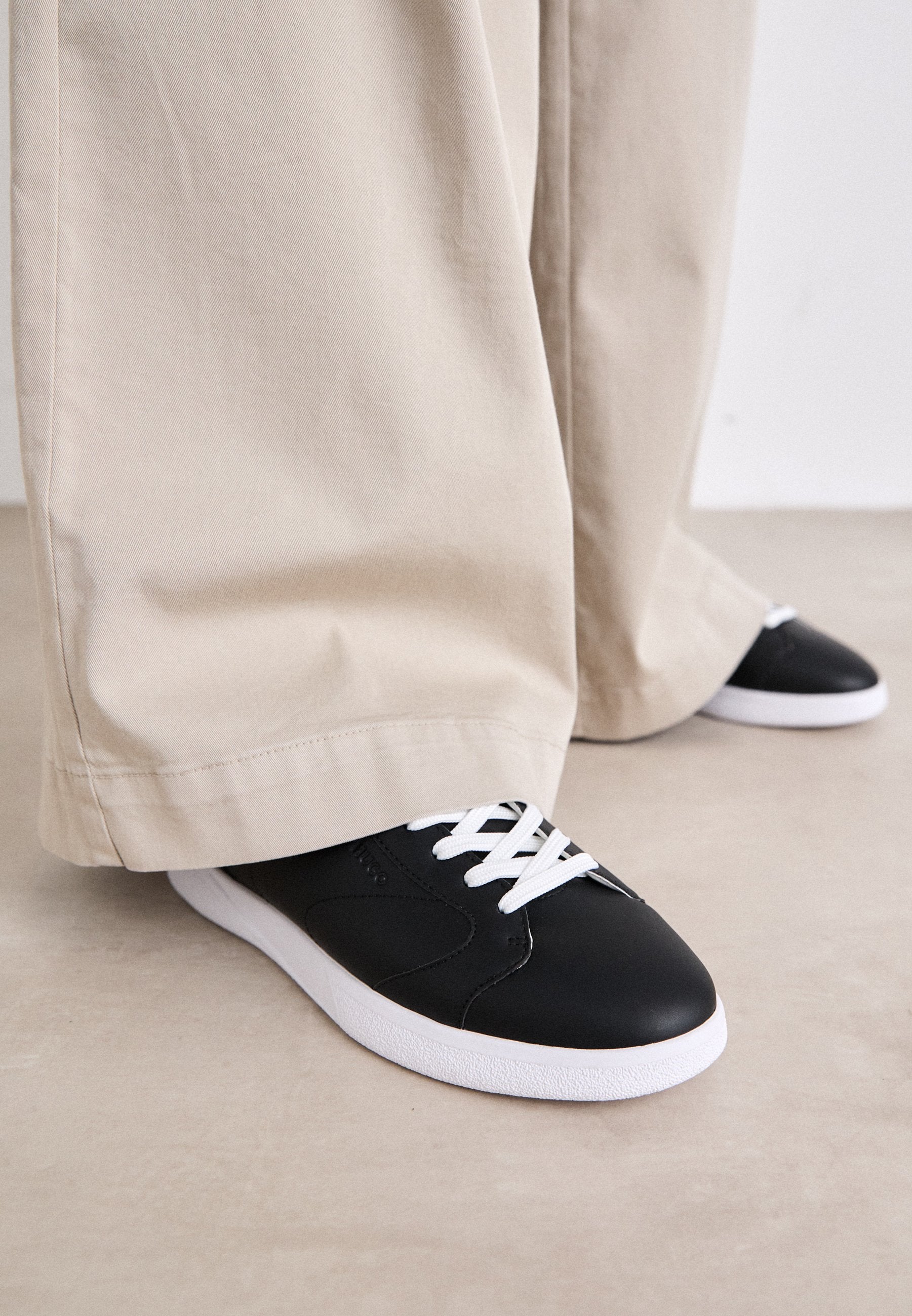 Schwarze Ledersneaker mit weißen Schnürsenkeln und einer weißen Gummisohle, kombiniert mit lockeren beigen Hosen, auf einer hellen strukturierten Oberfläche.