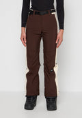 Person trägt braune und beige Skihose mit schwarzem Gürtel, schwarzen Stiefeln und schwarzem langarmigem Oberteil, steht vor einem schlichten Hintergrund.
