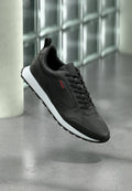 Schwarzer Ledersneaker mit weißer Sohle und schwarzen Schnürsenkeln, seitlich mit rotem "HUGO"-Logo, schwebt über einem grau polierten Boden.