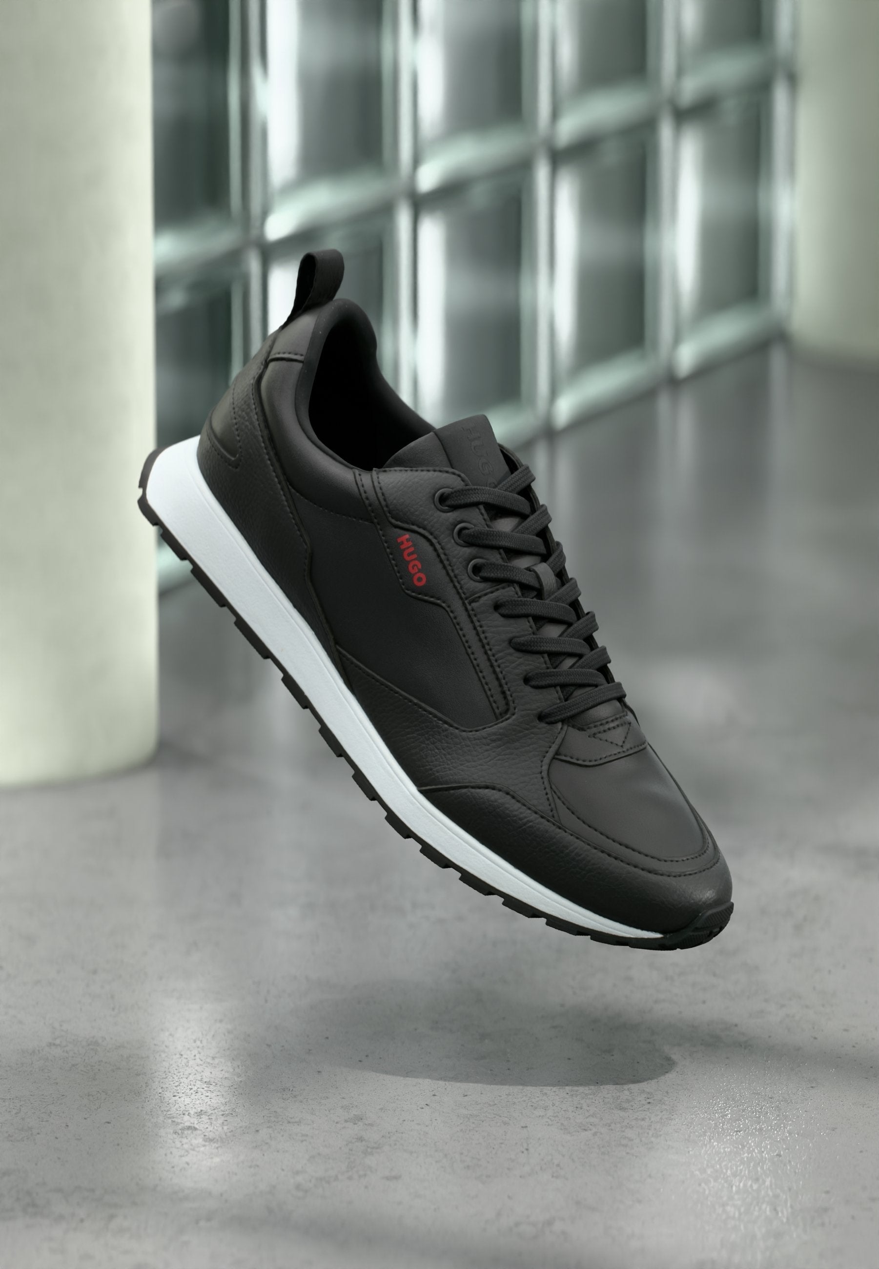 Schwarzer Ledersneaker mit weißer Sohle und schwarzen Schnürsenkeln, seitlich mit rotem "HUGO"-Logo, schwebt über einem grau polierten Boden.