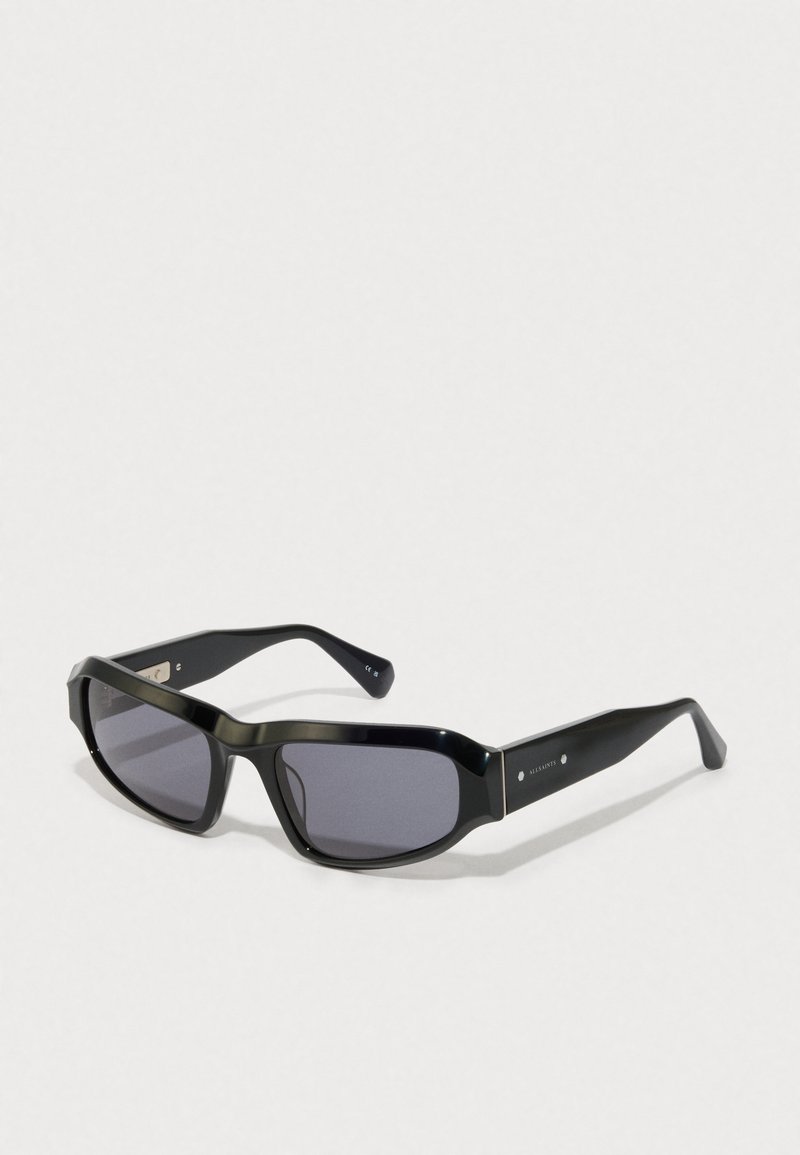 Schwarze rechteckige Sonnenbrille mit dunklen Gläsern und dicken Bügeln, mit einem kleinen silbernen Markenlogo an den Schläfen.