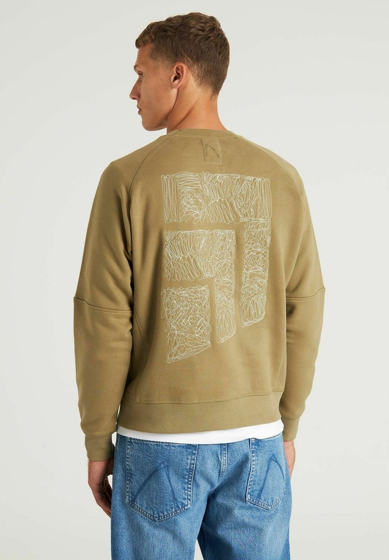 Olivgrüner Sweatshirt mit einer strukturierten weißen Grafik auf dem Rücken, langen Ärmeln und gerippten Bündchen. Über einem weißen Shirt getragen.