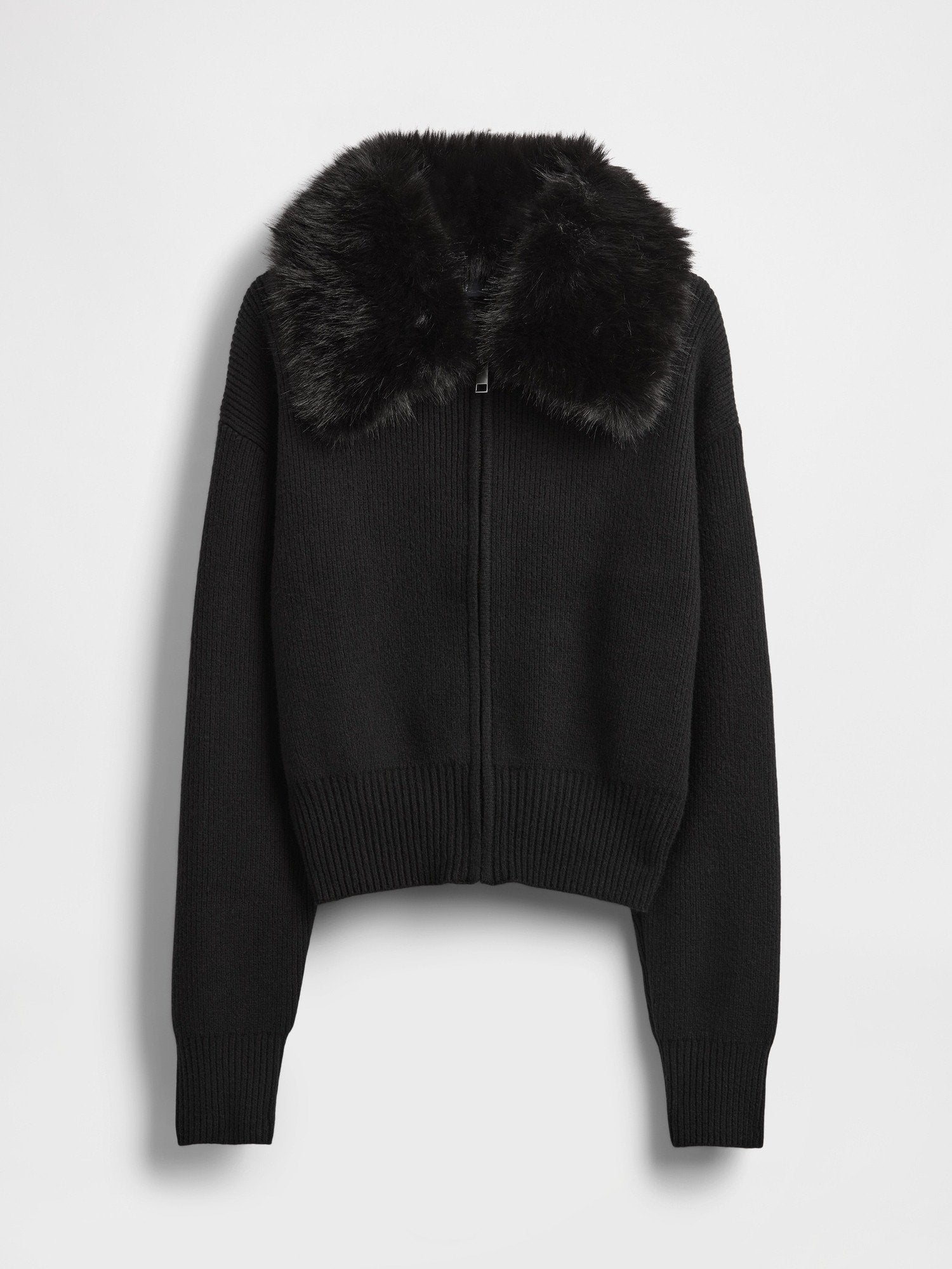 Schwarze, kurze Strickjacke mit gerippter Struktur, ausgestattet mit einem Kunstpelzkragen und einem Frontreißverschluss. Lange Ärmel mit enganliegenden Bündchen.