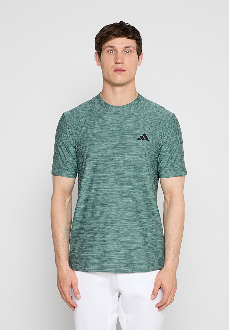 Mann, der frontal steht und ein grünes kurzärmeliges Adidas-T-Shirt sowie weiße Hose vor einem einfarbigen Hintergrund trägt.