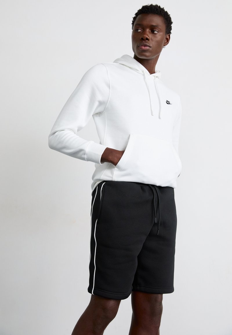 Junger Mann trägt einen weißen Nike-Hoodie mit Händen in der Fronttasche und schwarze Shorts mit weißen Seitenstreifen, steht vor einem einfarbigen Hintergrund.