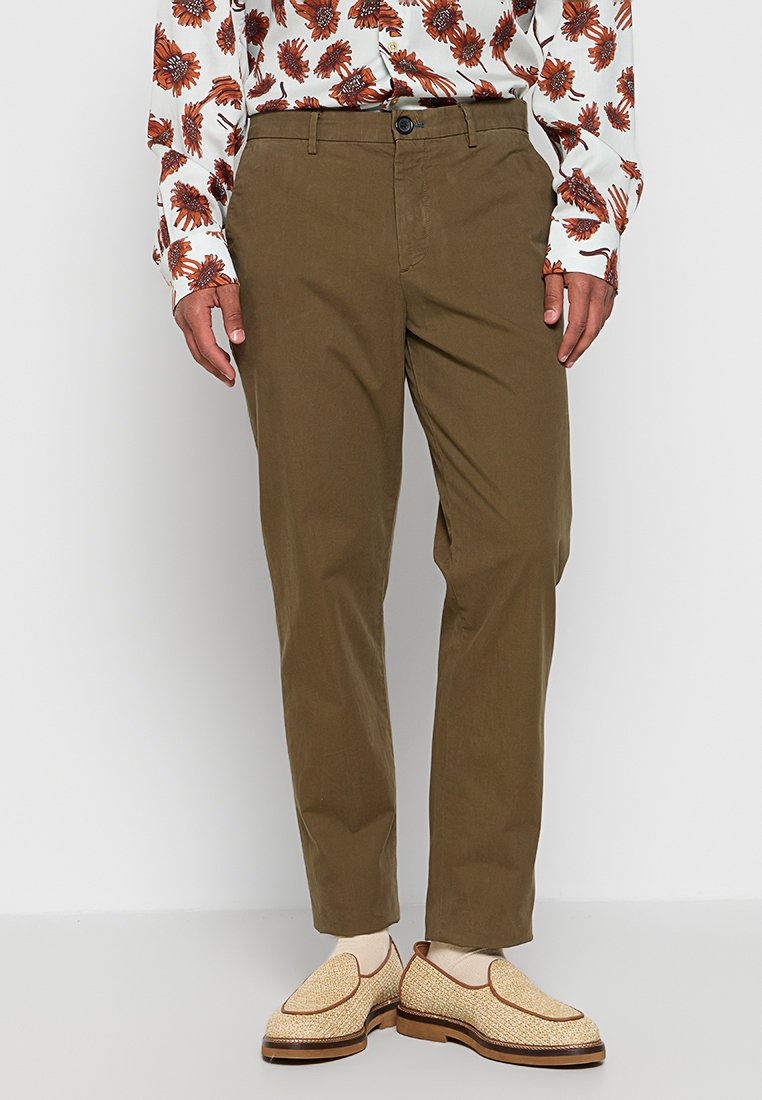 Eine Person steht da und trägt ein gemustertes Langarmhemd, olivgrüne Hosen und hellfarbige Loafers, was ein stilvolles Outfit präsentiert.