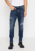 Mann in schmal geschnittenen, dunkelblauen, abgetragenen Jeans, schwarzen Knöchelstiefeln und einem weißen ärmellosen Oberteil, der vor einem weißen Hintergrund steht.