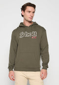 Junger Mann in einem olivgrünen Kapuzenpullover mit dem Schriftzug "Schott N.Y.C", beigen Hosen, steht vor einem schlichten hellen Hintergrund.