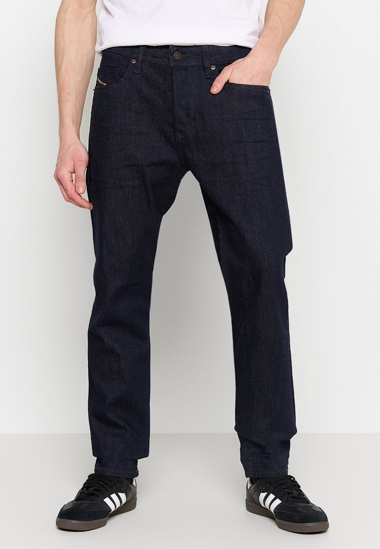 Dunkelblaue Jeans aus Denim mit geradem Schnitt, Fünf-Taschen-Design, vorderem Knopfverschluss und dezenten Nähten. Schwarze Sneakers mit weißen Streifen.