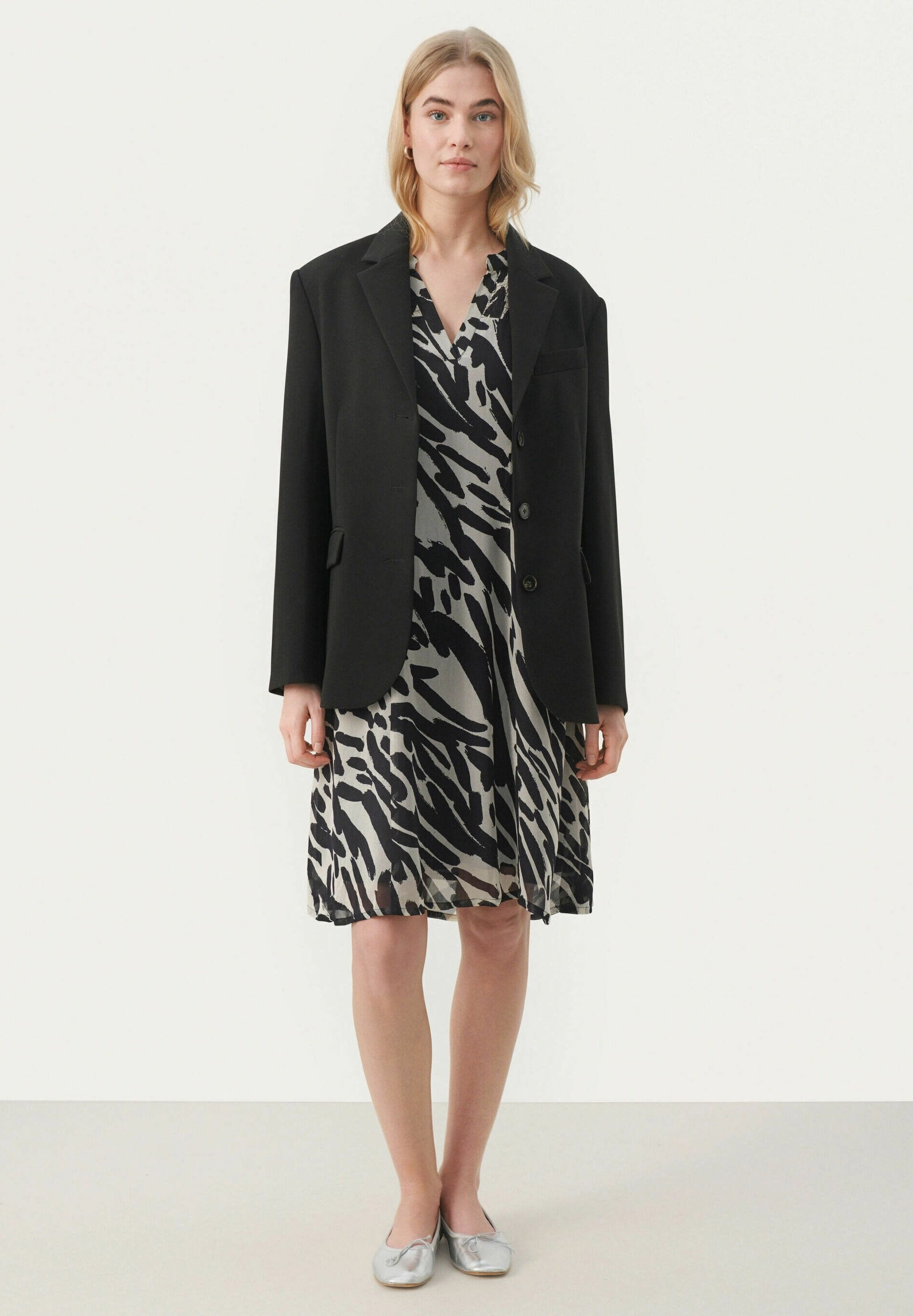 Schwarzer übergroßer Blazer über einem grauen Kleid mit einem schwarzen abstrakten Zebra-Print. Das Modell trägt silberne flache Schuhe. Einfaches, elegantes Design.