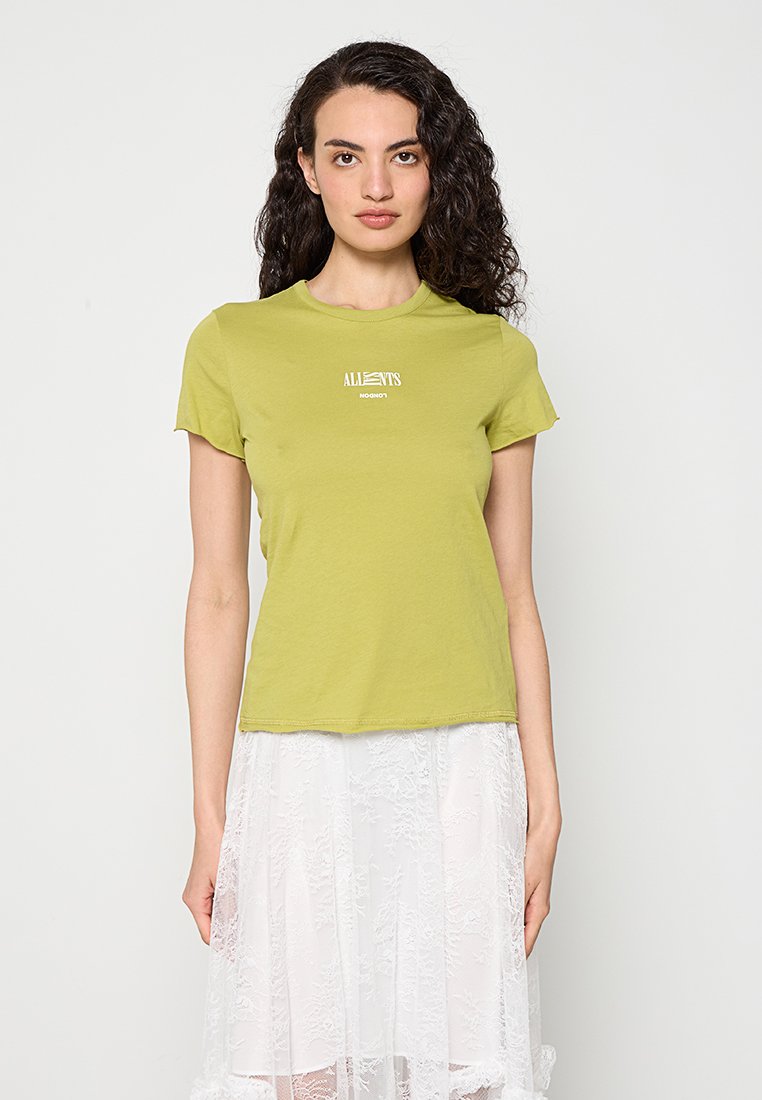 Frau mit lockigem dunklem Haar, die ein kurzärmeliges limettengrünes T-Shirt mit weißem Text und einen weißen Spitzenrock trägt, steht vor einem hellen Hintergrund.