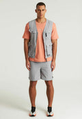 Graue Weste mit mehreren Taschen über einem pfirsichfarbenen T-Shirt, kombiniert mit hellgrauen Shorts und schwarzen Sneakers mit orangefarbenen Akzenten. Schlicher Hintergrund.