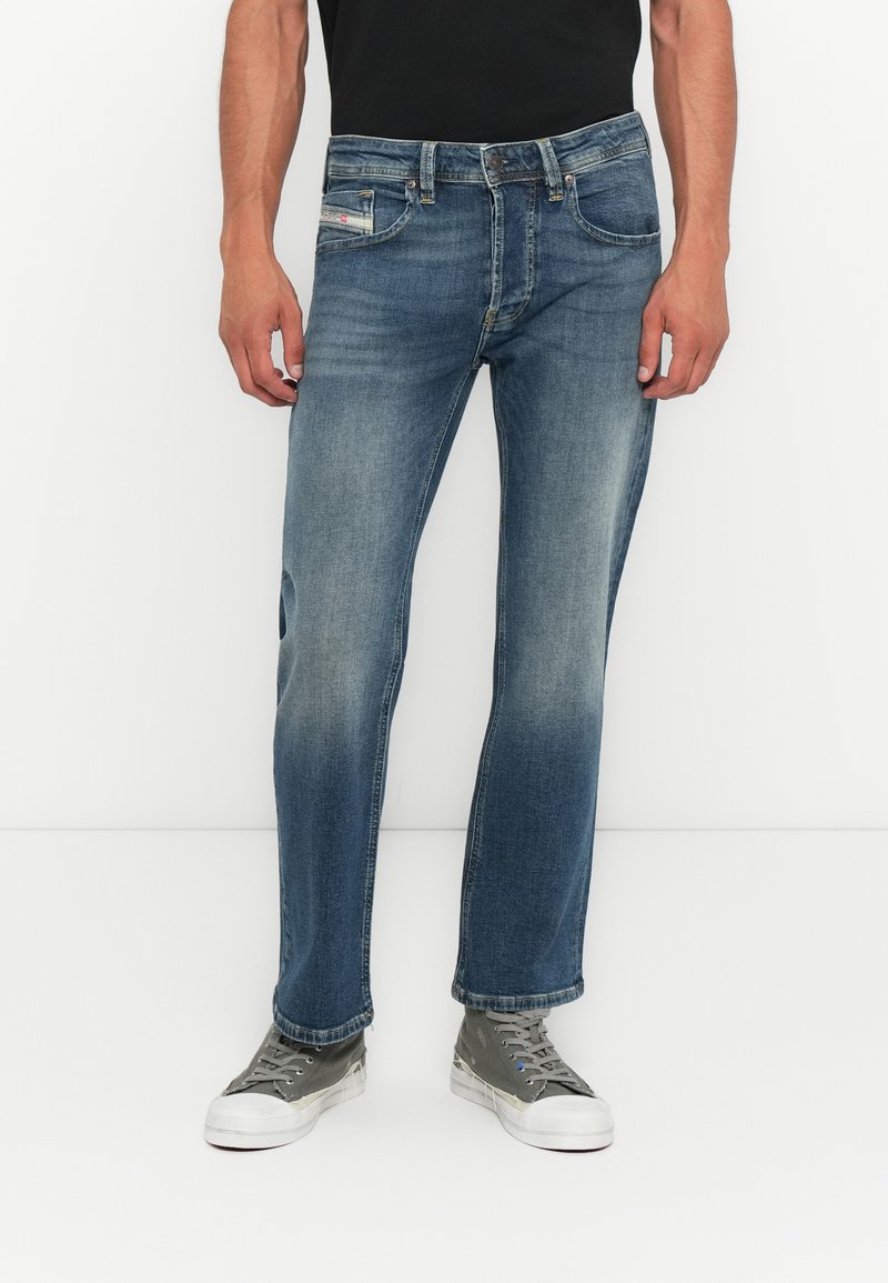 Mann in ausgewaschenen blauen Jeans und grauen Leinenschuhen, der gegen einen weißen Hintergrund steht, mit entspannten Händen an den Seiten.