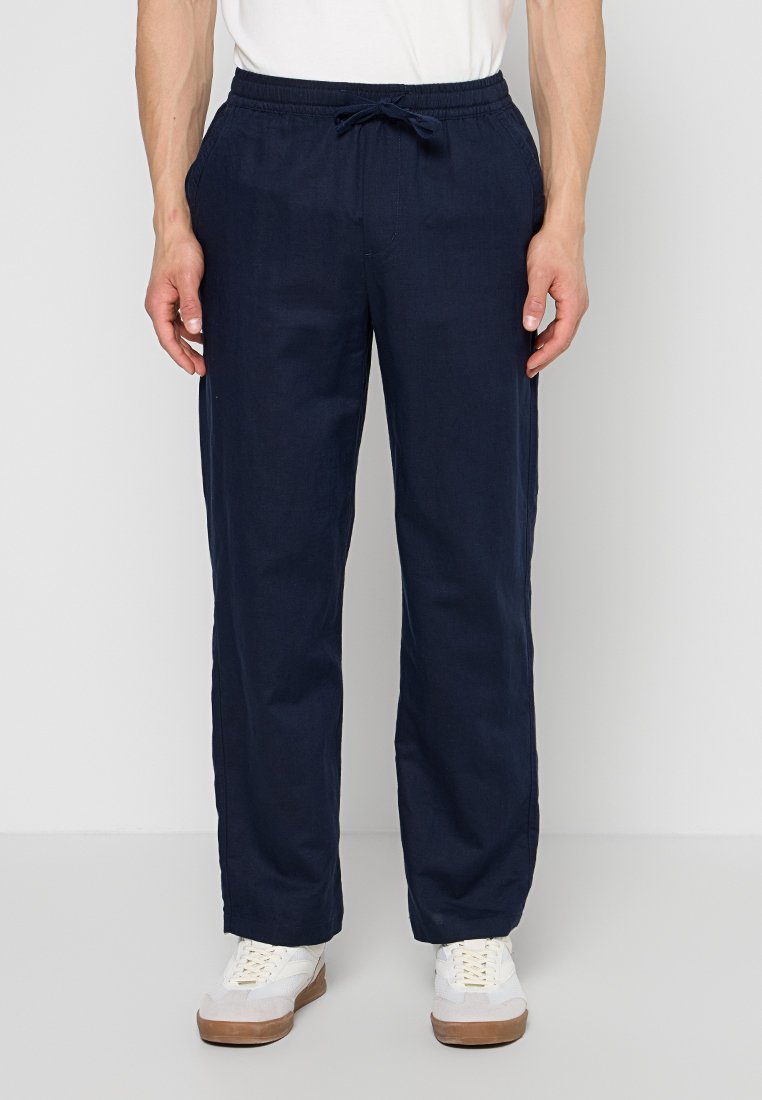 Mann trägt marineblaue Kordelhose und weiße Sneakers, steht vor einem einfarbig hellen Hintergrund.