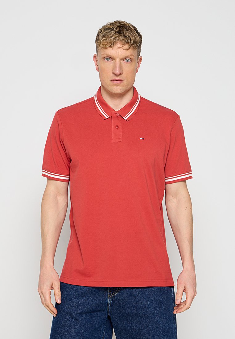 Mann trägt ein rotes Polohemd mit weiß gestreiftem Kragen und Ärmelsäumen, kombiniert mit dunkelblauen Jeans, steht vor einem einfarbigen Hintergrund.