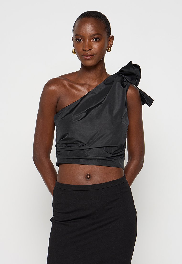 Frau mit kurzen Haaren, die goldene Ohrringe und ein schwarzes, einseitig geschnittenes Crop-Top mit einer Schleife trägt, kombiniert mit einem schwarzen Rock.